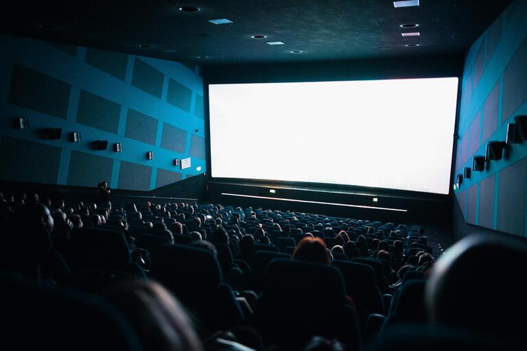 Photograph of a dimly lit movie theater with patrons sitting watching a white screen